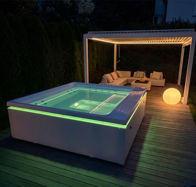 Ein moderner Whirlpool mit leuchtend grüner LED-Lichtleiste auf einem Holzdeck bei Nacht. Im Hintergrund eine beleuchtete Pergola mit Lounge-Möbeln.