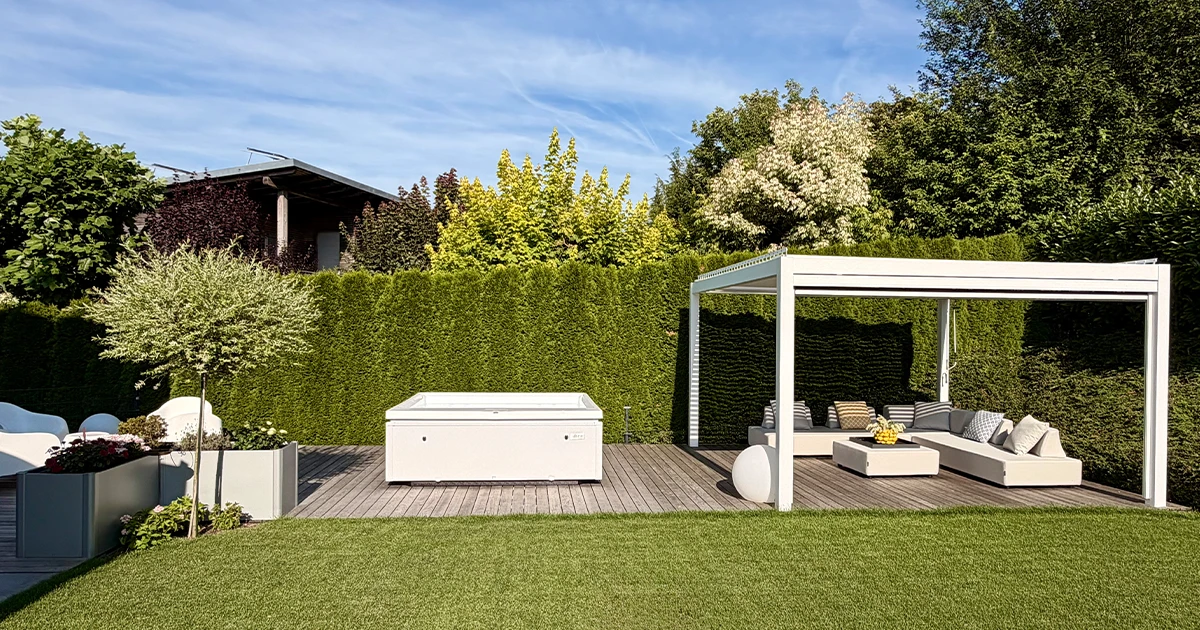 Eine Weitwinkel-Außenaufnahme eines modernen Luxus-Gartens bei Tageslicht. Der Whirlpool steht auf einem Holzdeck zwischen einem großen Pflanzkübel und einer Pergola mit Lounge-Sitzgruppe, umgeben von einer hohen Hecke.